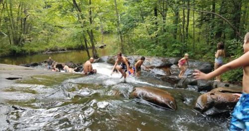 A group of children and adults play in a rocky stream surrounded by lush greenery. Water cascades over the rocks.