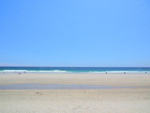A serene beach scene with gentle waves, clear blue skies, and a few people walking along the sandy shore.