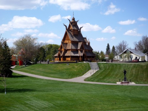 A wooden, ornate building with a steep roof stands on a grassy landscape under a blue sky with fluffy clouds.