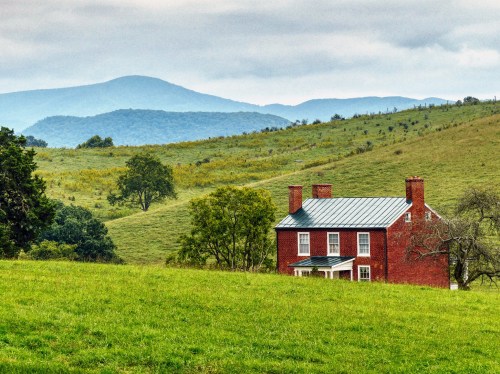 A red brick house sits in a green field, with rolling hills and mountains in the background under a cloudy sky.