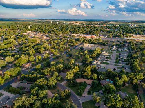 Aerial view of a suburban neighborhood with green trees, houses, and a distant skyline under a partly cloudy sky.