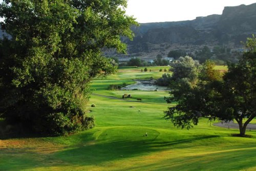Lush green golf course with trees, hills, and a serene water feature in the background under a clear sky.