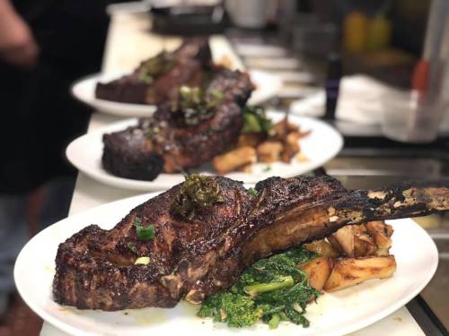 Three large, beautifully plated ribeye steaks with greens and roasted potatoes, served in a restaurant kitchen.