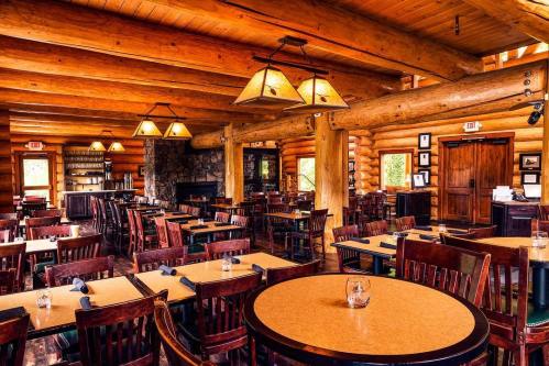 Cozy log cabin restaurant interior with wooden tables, chairs, and warm lighting. Stone fireplace in the background.