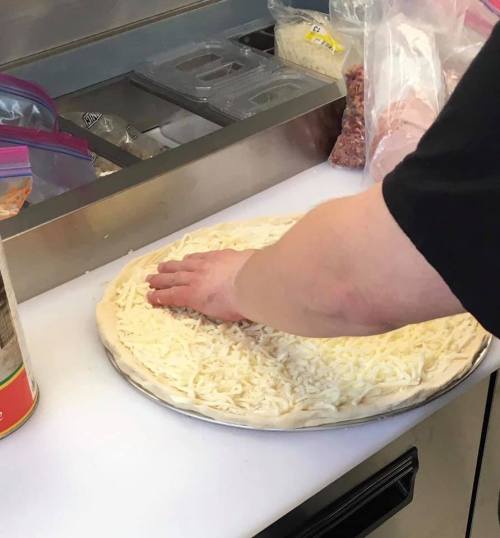 A hand pressing shredded cheese onto a pizza crust on a countertop, with ingredients in the background.