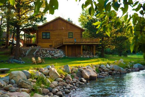 A rustic log cabin by a serene river, surrounded by trees and large rocks, with a wooden deck and chairs.