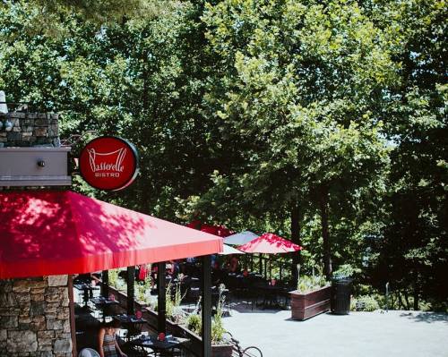 A bistro with a red awning and outdoor seating, surrounded by lush green trees.