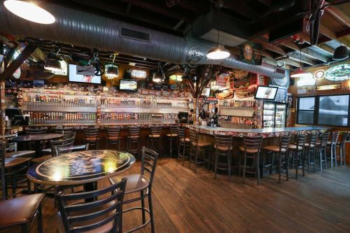 A cozy bar interior with a long beer tap wall, wooden tables, and chairs, featuring various beer signs and decor.