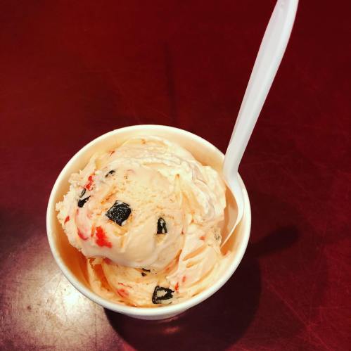 A cup of ice cream with swirls of orange and black, topped with chunks of chocolate and a white spoon.