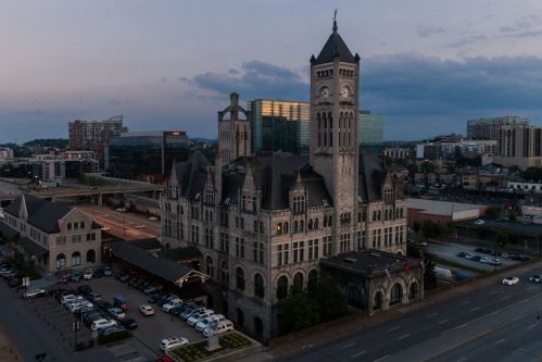 Historic building with a clock tower, surrounded by modern architecture and parked cars at dusk.