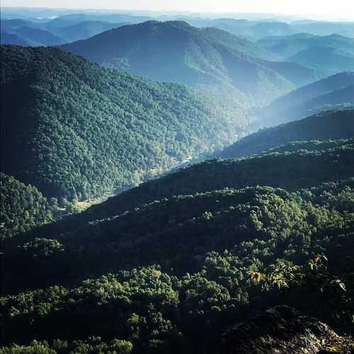A panoramic view of lush green mountains and valleys under a clear sky, with soft sunlight illuminating the landscape.