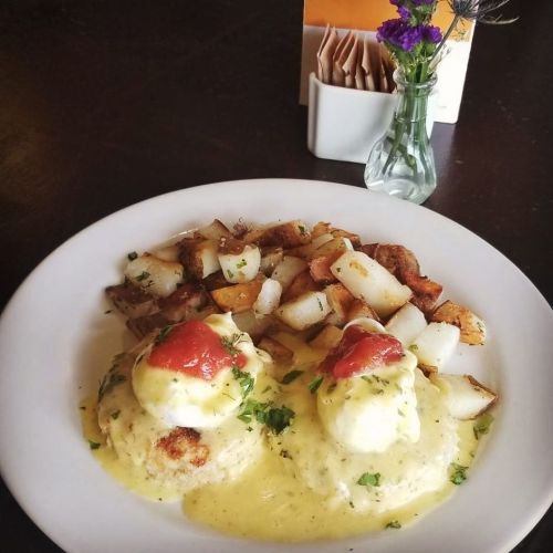 A plate of eggs Benedict with hollandaise sauce, topped with tomato, served with seasoned potato cubes and a small flower vase.