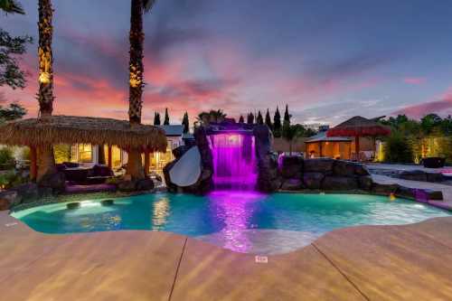 A vibrant pool with a waterfall and slide, surrounded by palm trees and tiki huts, under a colorful sunset sky.