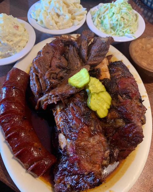 A hearty plate of barbecue featuring ribs, sausage, pickles, and sides of coleslaw and macaroni salad.