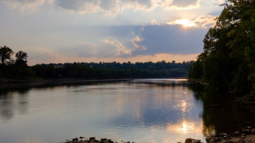 A serene river scene at sunset, with clouds reflecting on the water and trees lining the shore.