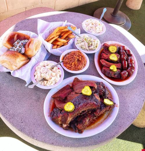 A table spread with barbecue ribs, sausage, sides of coleslaw, baked beans, potato salad, fries, and bread rolls.