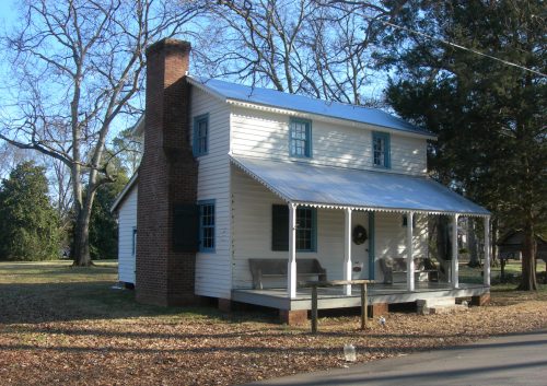 A white, two-story house with a porch, green shutters, and a chimney, surrounded by trees and fallen leaves.