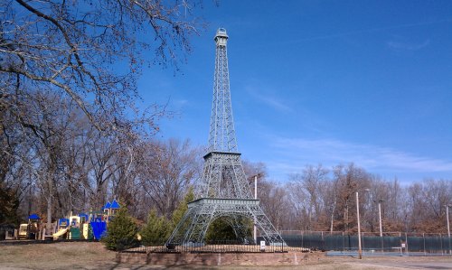 A miniature Eiffel Tower stands in a park, surrounded by trees and a playground in the background under a clear blue sky.
