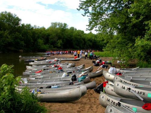 Canoes lined up on a sandy shore with a group of people gathered by the riverbank surrounded by lush greenery.