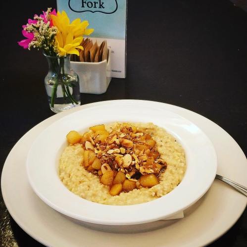 A bowl of oatmeal topped with caramelized apples and almonds, served with a small vase of flowers in the background.