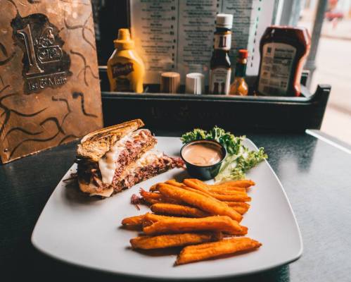 A hearty sandwich with layers of meat and sauce, served with sweet potato fries and a side of dipping sauce.