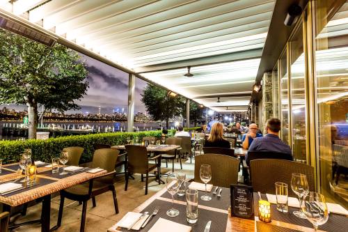 A stylish restaurant patio with tables set for dining, overlooking a city skyline at night.