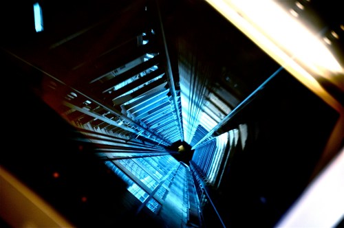 A downward view of an elevator shaft, showcasing metal beams and a blue-lit interior.