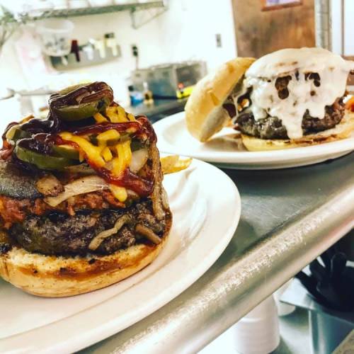 Two gourmet burgers on plates: one topped with colorful vegetables and sauces, the other with melted cheese.