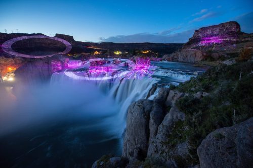 A waterfall illuminated by colorful lights at dusk, surrounded by rocky cliffs and a serene river.