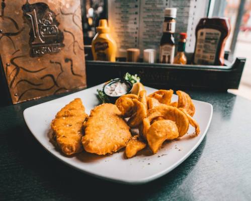 A plate of golden fried fish fillets with curly fries and a side of dipping sauce, set on a dark table.