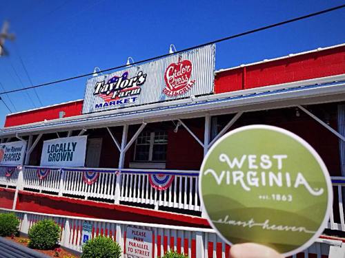 A person holds a "West Virginia" sticker in front of Taylor's Farm Market, featuring a red building and local produce signs.