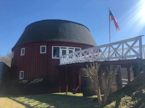Red round barn with a white railing, American flag flying, set against a clear blue sky.