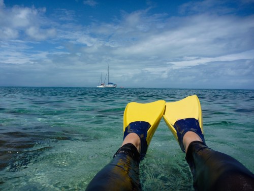 Person's feet in yellow flippers submerged in clear water, with a sailboat in the distance under a partly cloudy sky.