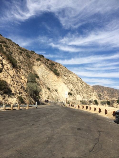 Winding road alongside a rocky hillside under a blue sky with wispy clouds. Road signs visible.