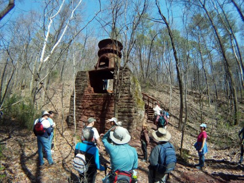 A group of hikers gathers around a stone structure in a wooded area, surrounded by trees and fallen leaves.