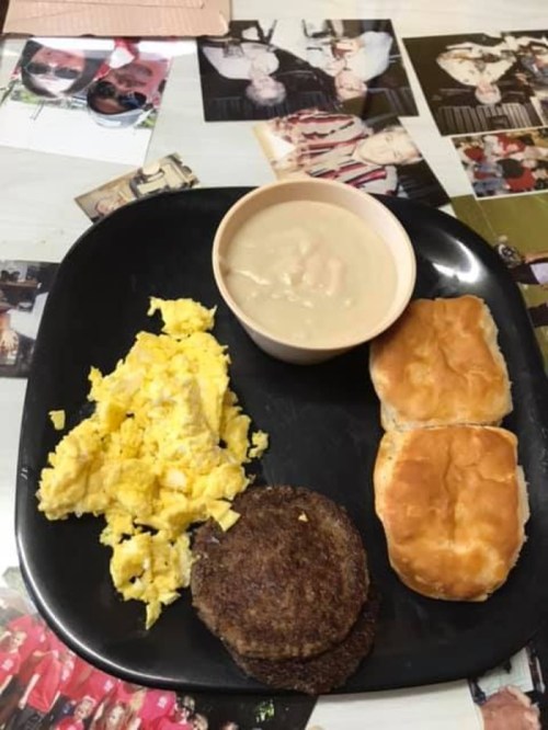 A black plate with scrambled eggs, two biscuits, a bowl of soup, and two sausage patties on a table with photos.