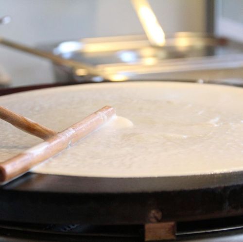 A close-up of a cooking surface with a wooden spatula spreading batter for a crepe or pancake.