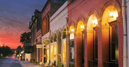Historic buildings line a quiet street at dusk, illuminated by warm lights under a colorful sky.