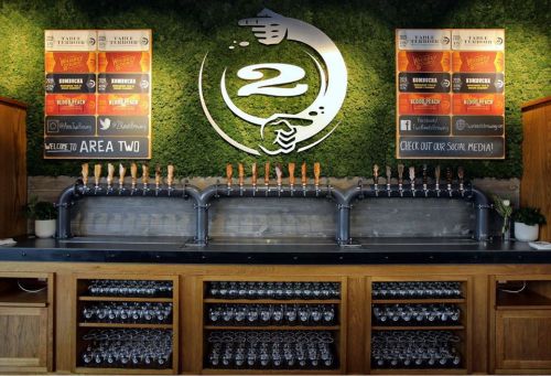 A bar with a green moss wall, featuring a logo, beer taps, and a display of glasses underneath.