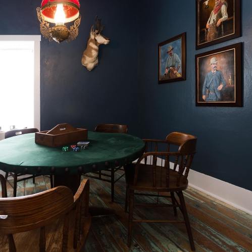 A cozy room with a green table, wooden chairs, and framed portraits on a dark blue wall. A mounted deer head is visible.