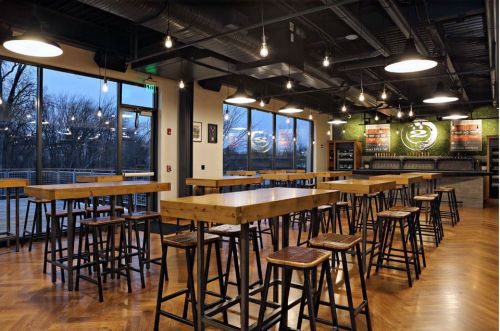 Modern bar interior with wooden tables and stools, large windows, and a well-lit atmosphere.