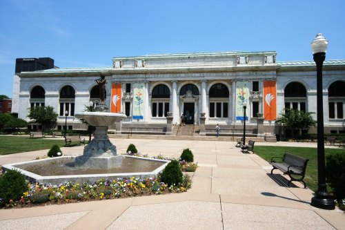 A grand library building with a fountain and flower beds in the foreground, under a clear blue sky.