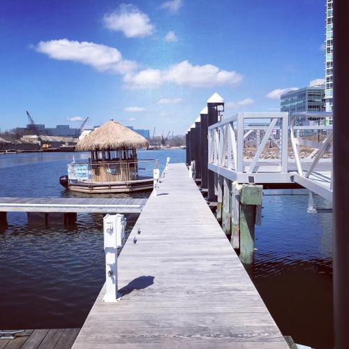 A wooden dock extends over water, leading to a thatched-roof hut under a clear blue sky with scattered clouds.