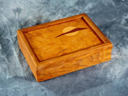 A wooden box with a smooth finish and a circular design on the lid, resting on a textured gray surface.