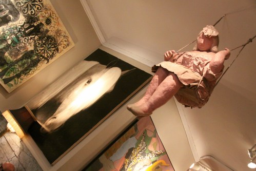 A suspended sculpture of a seated figure in a pink dress, with abstract paintings and a horse portrait in the background.