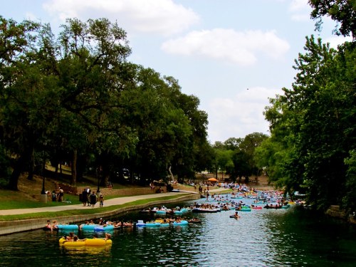 A busy river scene with people floating on colorful tubes, surrounded by trees and a grassy bank under a blue sky.