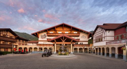 A charming alpine-style hotel with a grand entrance, surrounded by mountains and a colorful sky at sunset.