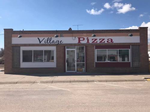 A brick building with a sign reading "Village Pizza" and large windows, set against a blue sky.