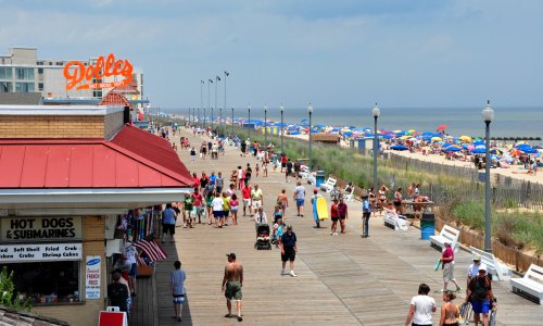 A busy boardwalk with people walking, shops, and colorful umbrellas on the beach in the background.