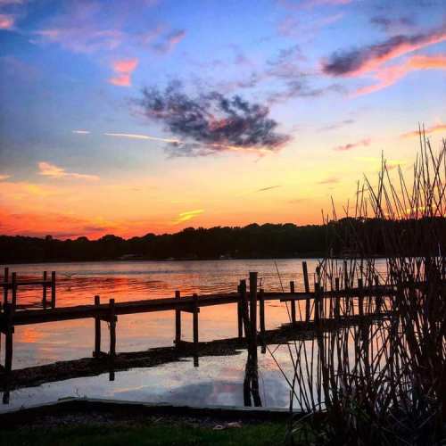 A serene sunset over a lake, with colorful clouds reflecting on the water and silhouettes of wooden docks and reeds.